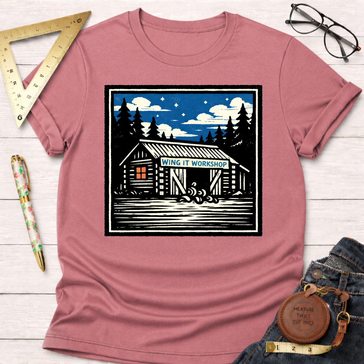 Heather dusty rose cotton tee with "Wing It Workshop" cabin graphic, styled with jeans and tools.