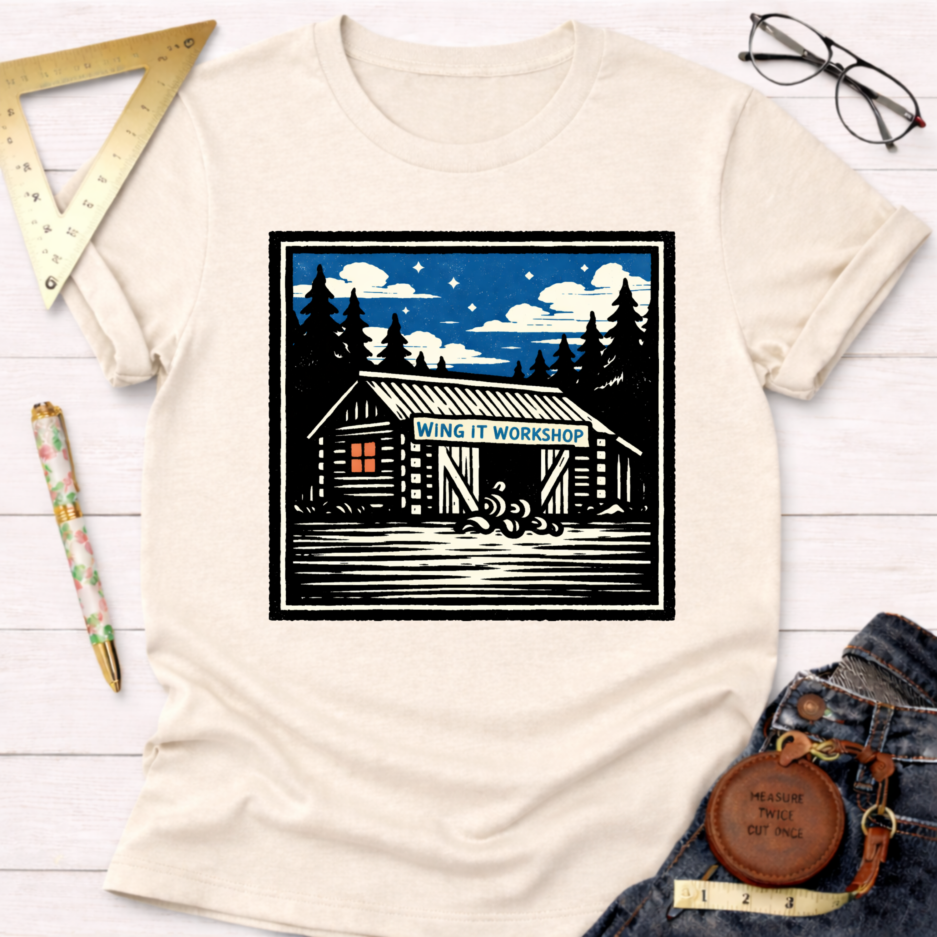 Cream "Wing It Workshop" graphic tee with a cabin and pine trees design, styled with jeans and tools.