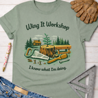 Women's funny woodworking graphic tee shirt for female woodworkers, makers, and DIY builders — Sawdust & Ponytails. Light green cotton tee with "Wing It Workshop" and "I know what I'm doing" text, featuring a saw, screwdriver, stump, and cabin graphic.