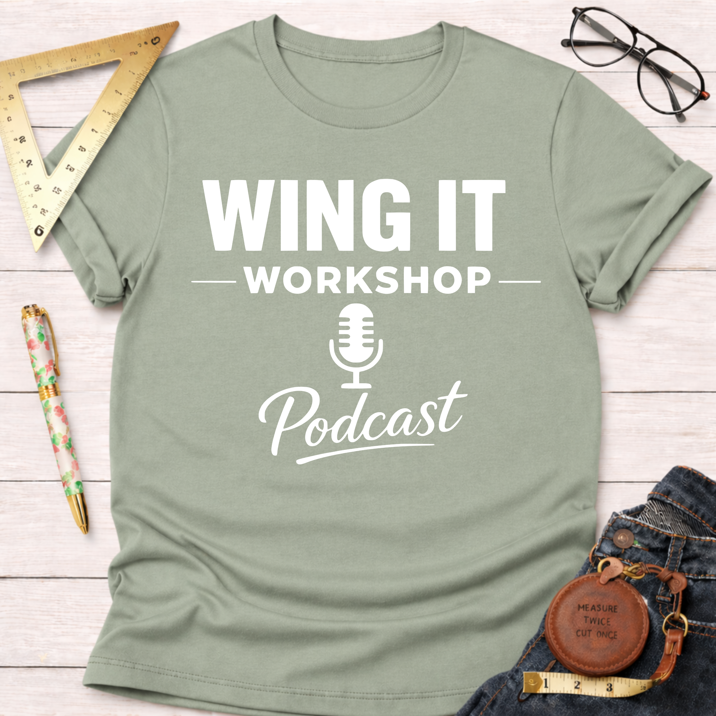 Sage green cotton "Wing It Workshop Podcast" t-shirt with white text, flat lay on a wooden surface.