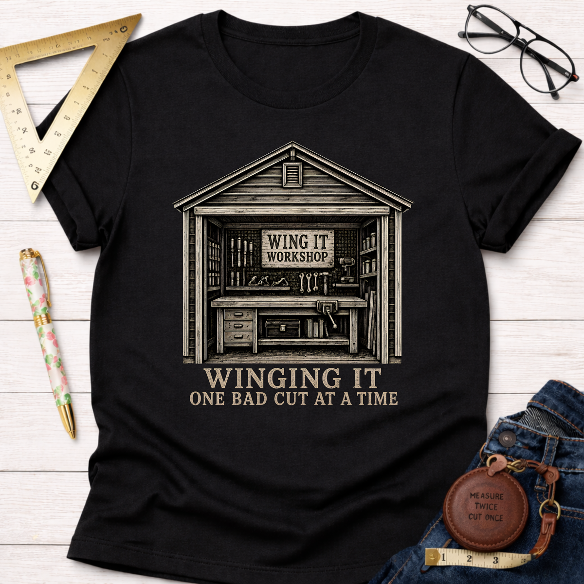 Black t-shirt with "Winging It Workshop" design featuring a shed full of tools and "One Bad Cut At A Time" text.