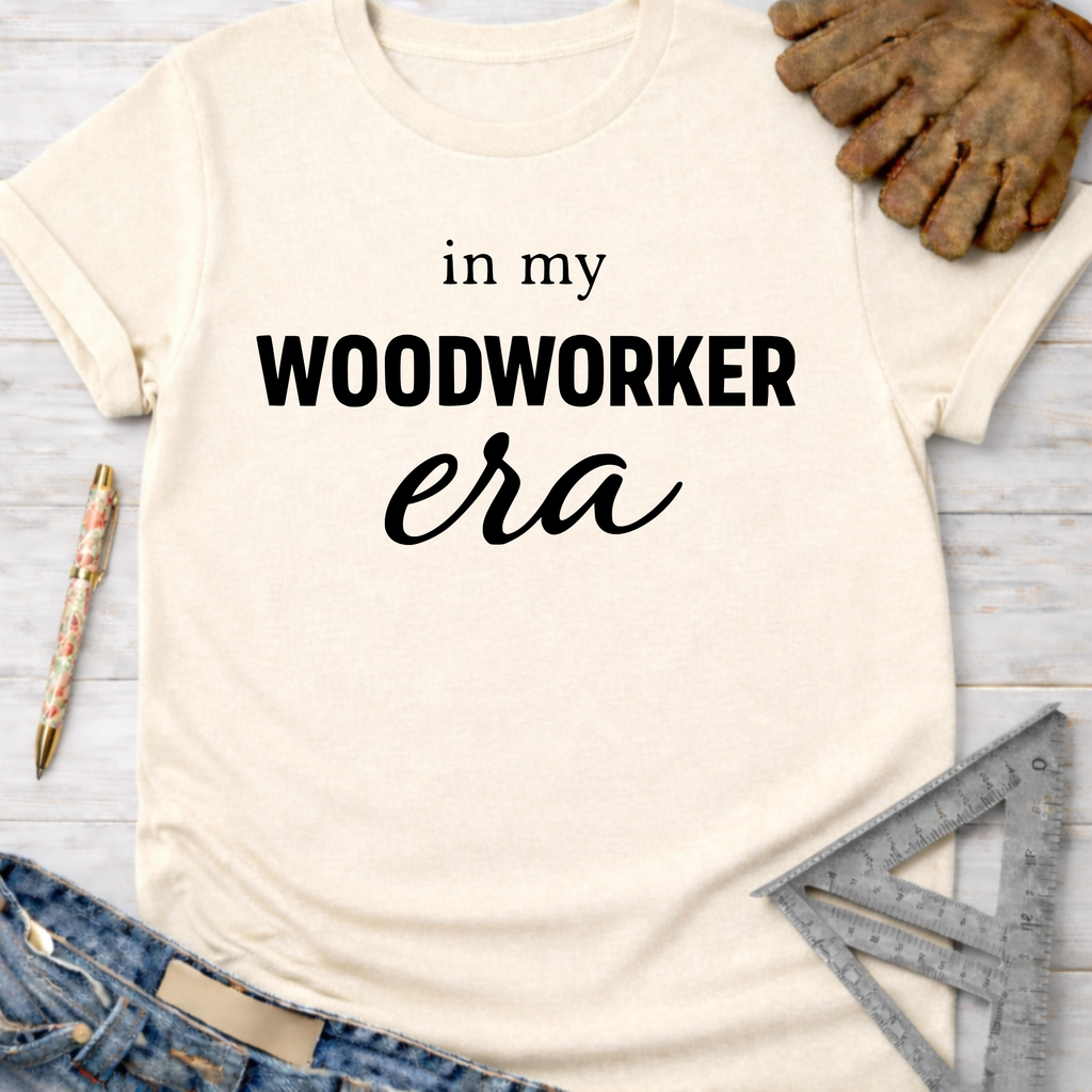 Women's funny woodworking graphic tee shirt for female woodworkers, makers, and DIY builders — Sawdust & Ponytails. Cream short-sleeve t-shirt with "in my WOODWORKER era" text, jeans, work glove, pen, and ruler.