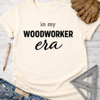 Women's funny woodworking graphic tee shirt for female woodworkers, makers, and DIY builders — Sawdust & Ponytails. Cream short-sleeve t-shirt with "in my WOODWORKER era" text, jeans, work glove, pen, and ruler.