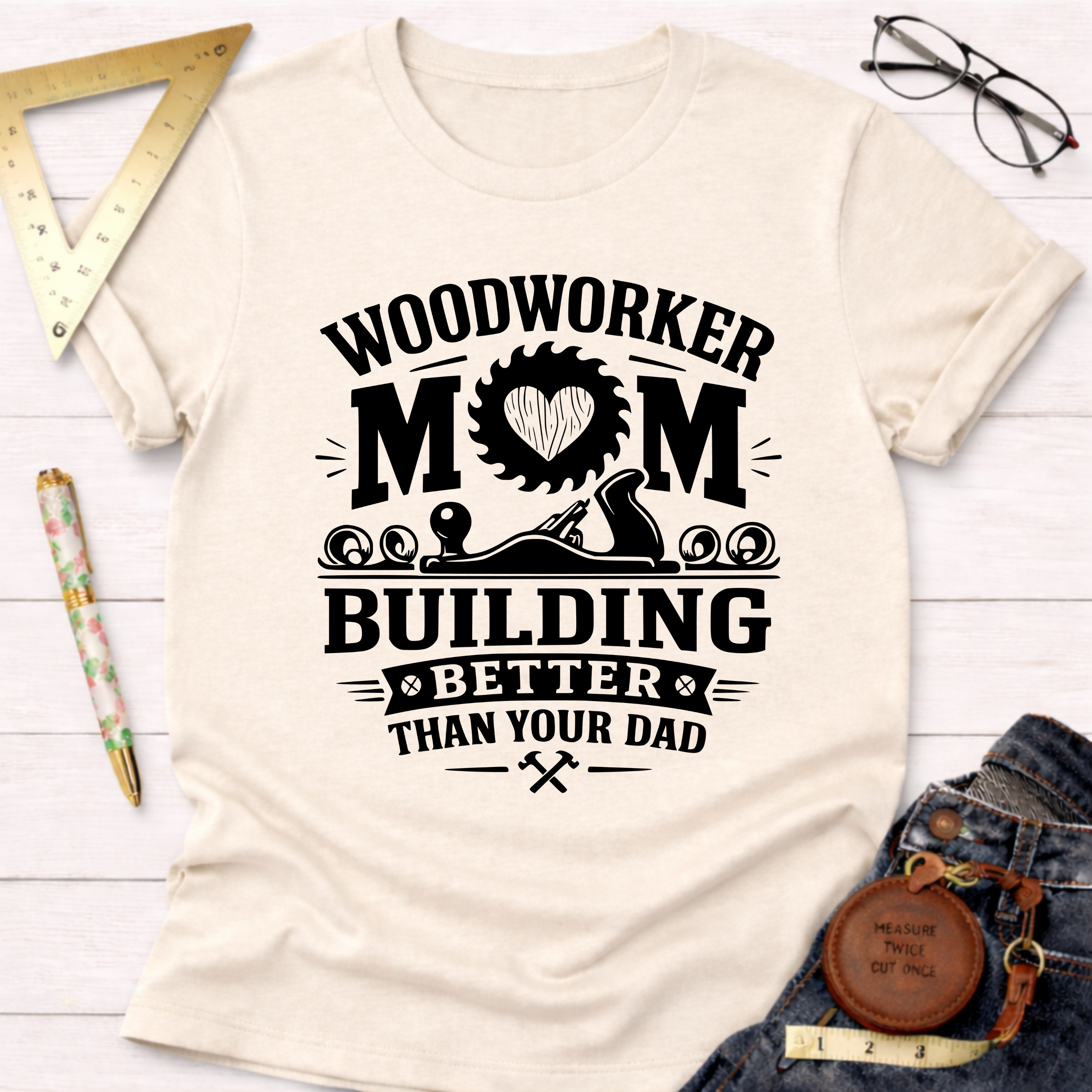 Women's funny woodworking graphic tee shirt for female woodworkers, makers, and DIY builders — Sawdust & Ponytails. Cream t-shirt with "Woodworker Mom Building Better Than Your Dad" graphic.