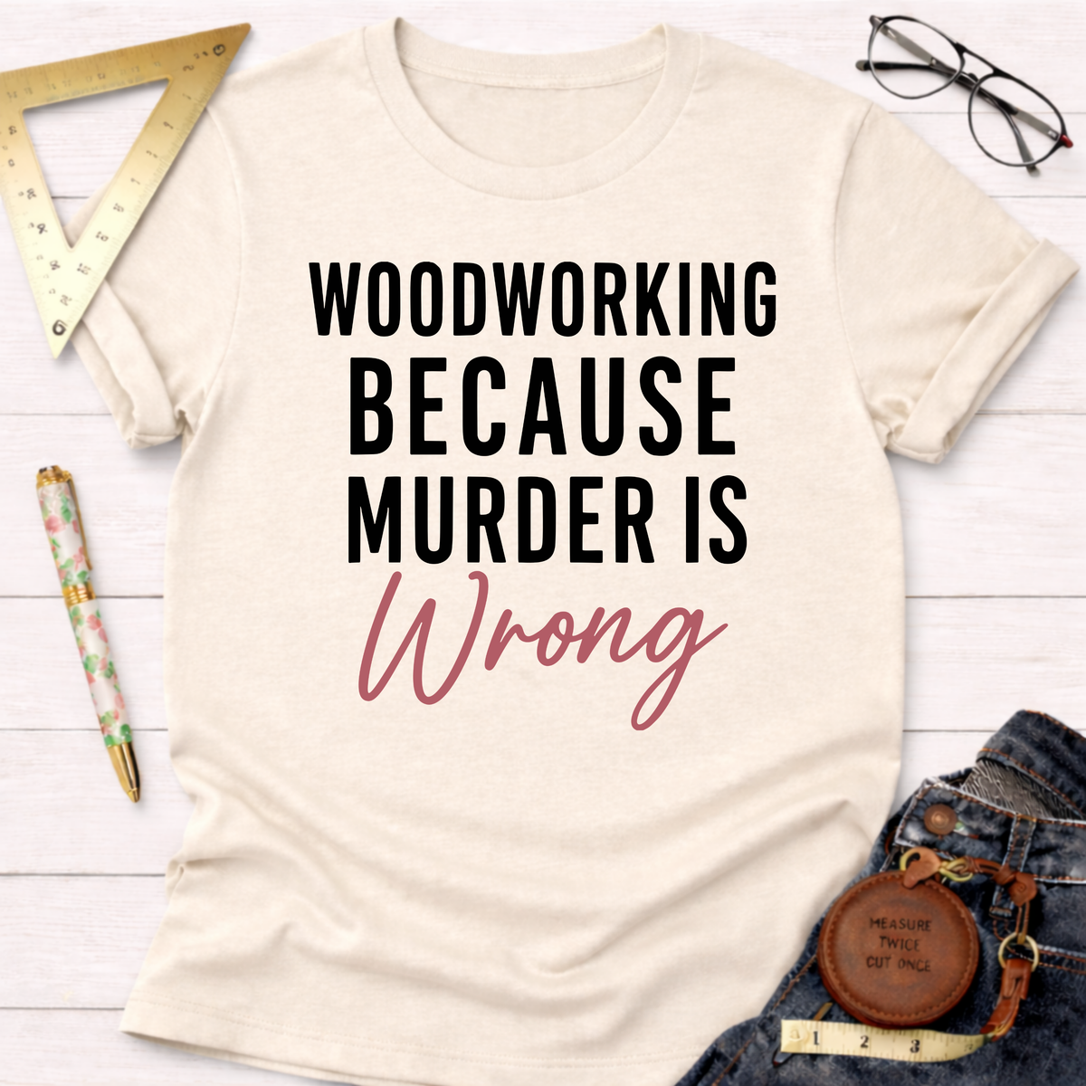 Cream t-shirt with "WOODWORKING BECAUSE MURDER IS Wrong" text in black and pink, styled with jeans and tools.