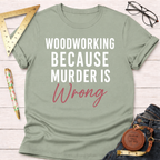 Sage green t-shirt with "WOODWORKING BECAUSE MURDER IS Wrong" text in white and pink, folded on a wooden surface.