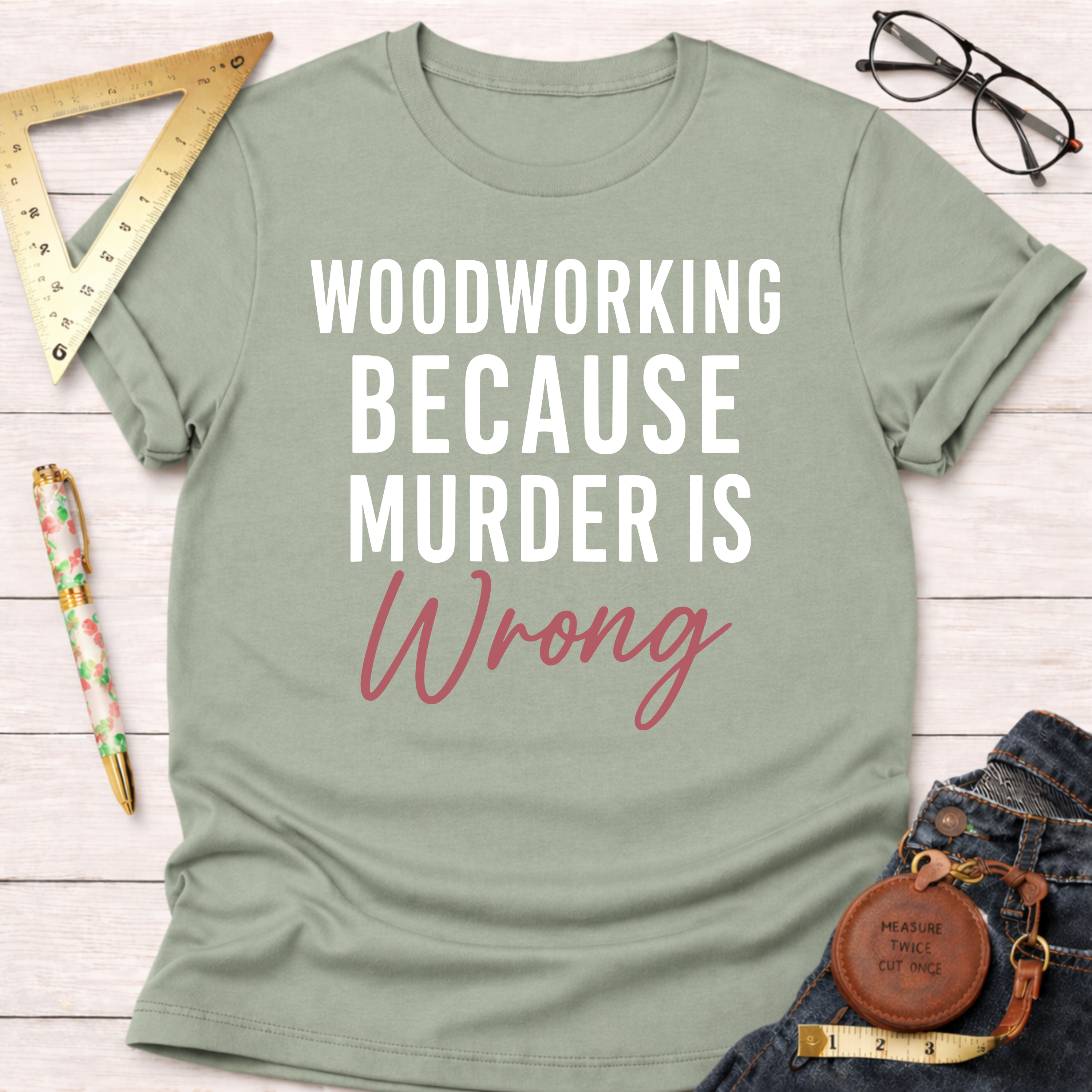 Sage green t-shirt with "WOODWORKING BECAUSE MURDER IS Wrong" text in white and pink, folded on a wooden surface.