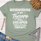 Women's funny woodworking graphic tee shirt for female woodworkers, makers, and DIY builders — Sawdust & Ponytails. Sage green cotton t-shirt with "Woodworking Is My Therapy Still Cheaper Than Actual Therapy" in white text.