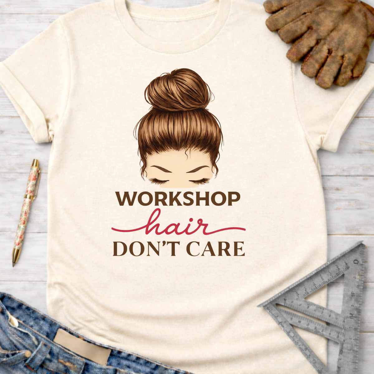 Women's funny woodworking graphic tee shirt for female woodworkers, makers, and DIY builders — Sawdust & Ponytails. Cream cotton tee with "Workshop Hair Don't Care" graphic and messy bun illustration.