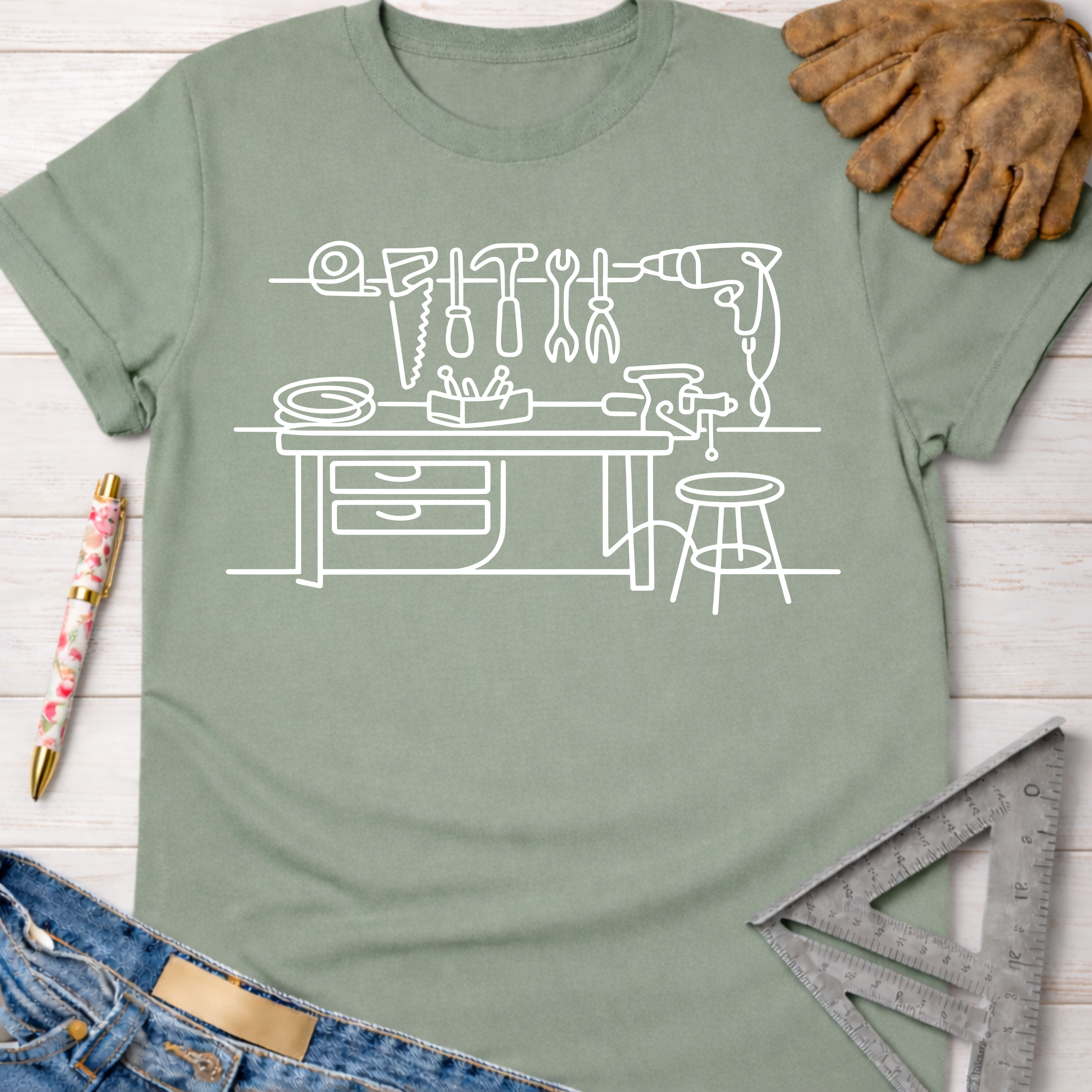 Women's funny woodworking graphic tee shirt for female woodworkers, makers, and DIY builders — Sawdust & Ponytails. Sage green t-shirt with white line art of a workshop bench and tools.