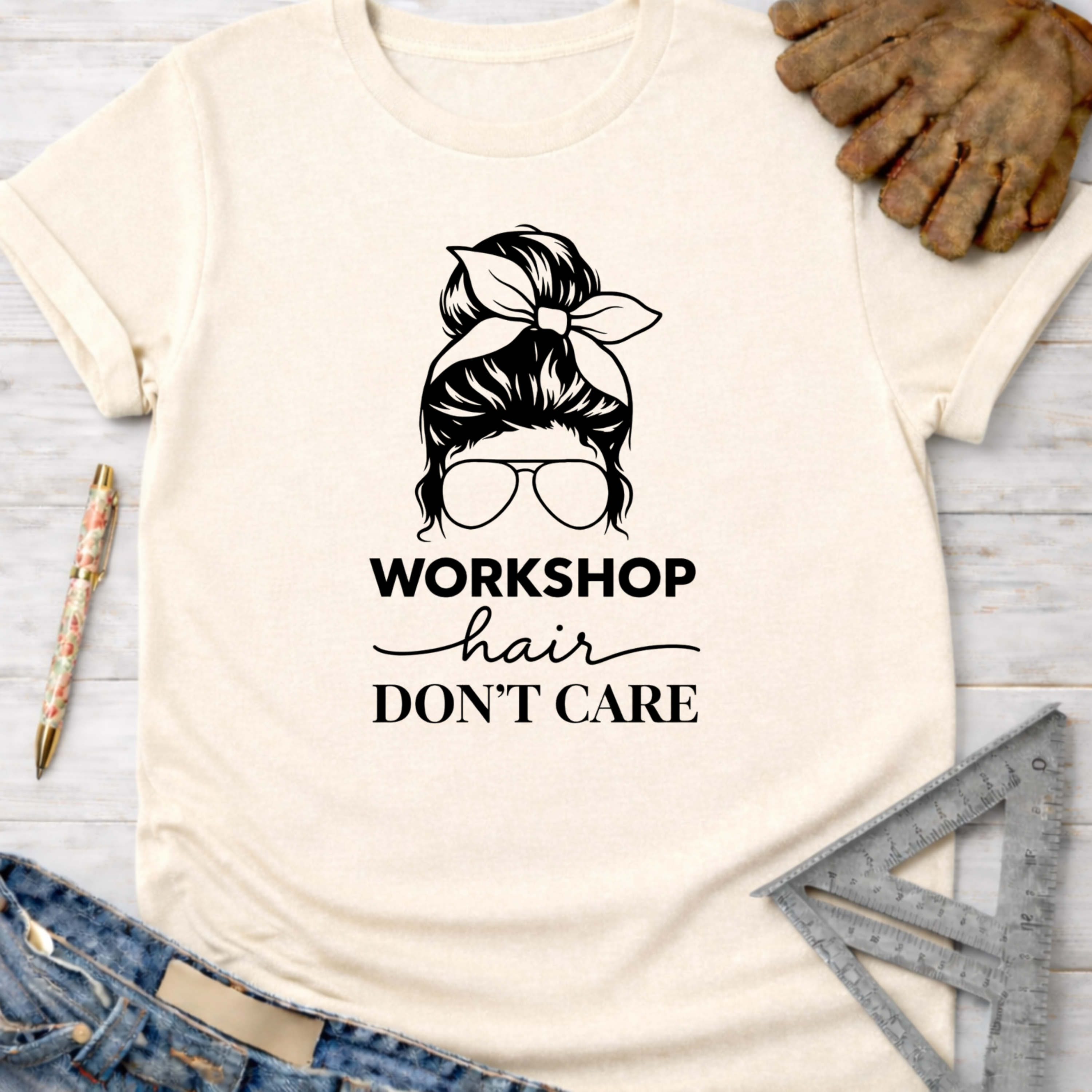 Women's funny woodworking graphic tee shirt for female woodworkers, makers, and DIY builders — Sawdust & Ponytails. Cream cotton tee with "Workshop Hair Don't Care" graphic of a messy bun and sunglasses.
