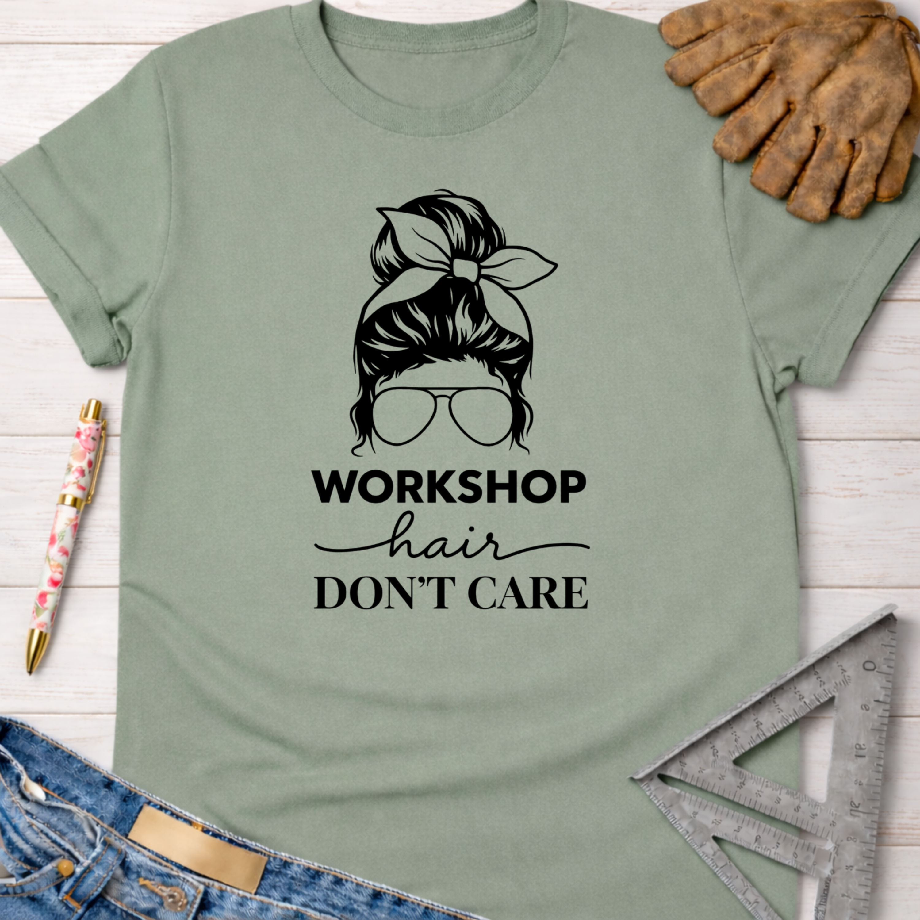 Women's funny woodworking graphic tee shirt for female woodworkers, makers, and DIY builders — Sawdust & Ponytails. Sage green cotton t-shirt with "Workshop Hair Don't Care" graphic of a messy bun and sunglasses.