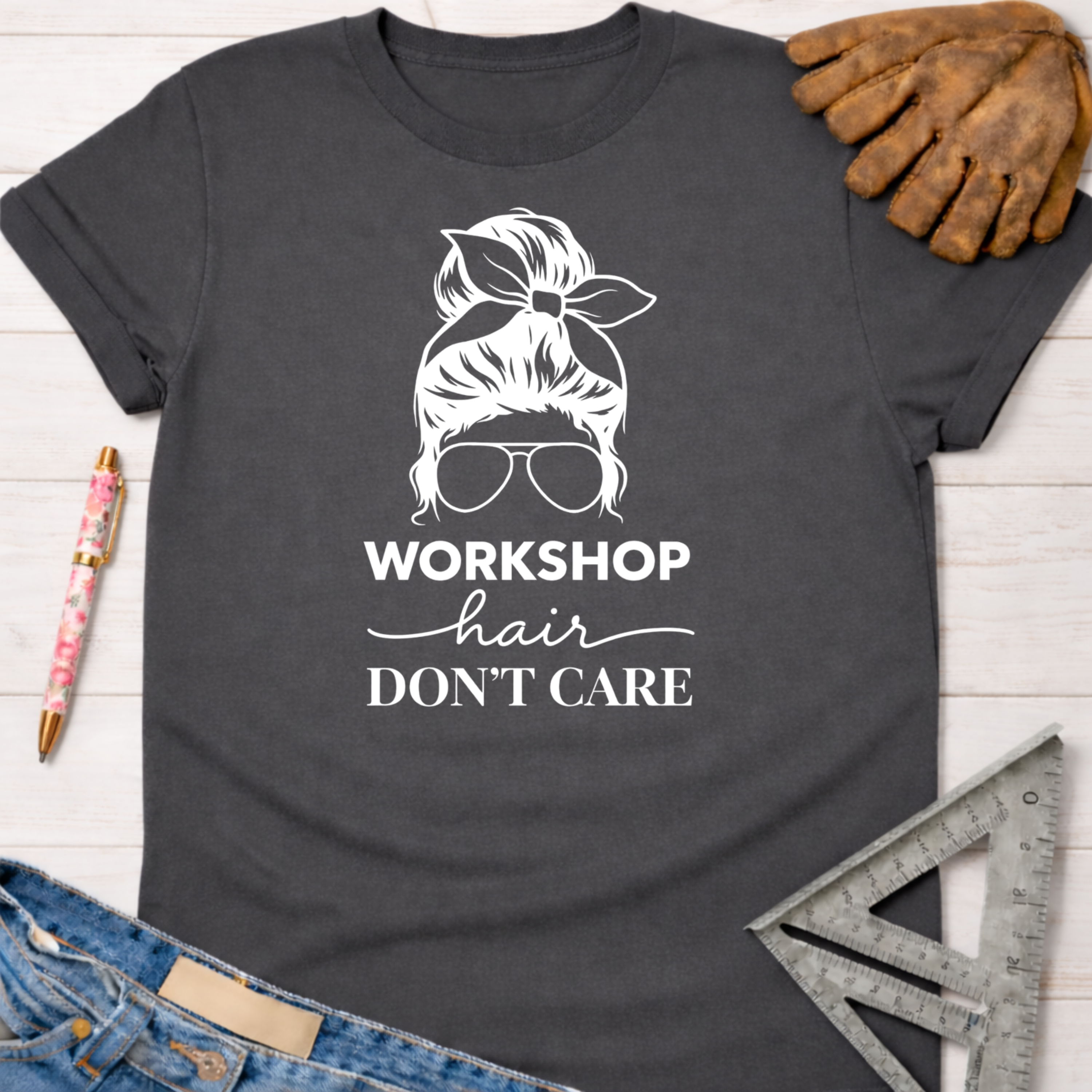 Women's funny woodworking graphic tee shirt for female woodworkers, makers, and DIY builders — Sawdust & Ponytails. Dark gray t-shirt with "Workshop Hair Don't Care" graphic, messy bun, and sunglasses.