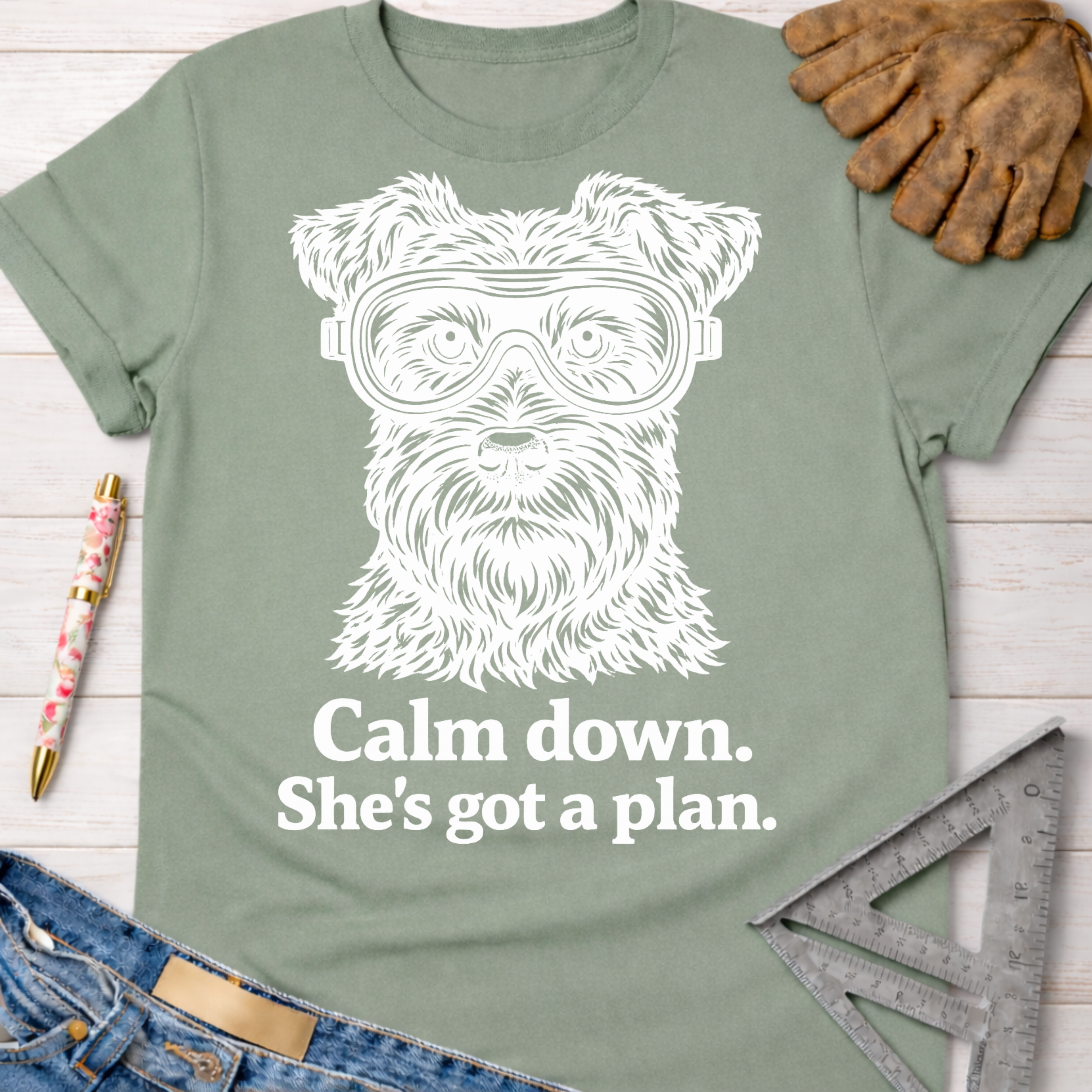 Women's funny woodworking graphic tee shirt for female woodworkers, makers, and DIY builders — Sawdust & Ponytails. Sage green cotton t-shirt with a white graphic of a dog wearing safety goggles and the text "Calm down. She's got a plan.