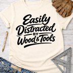 Women's funny woodworking graphic tee shirt for female woodworkers, makers, and DIY builders — Sawdust & Ponytails. Cream short-sleeve tee with "Easily Distracted by Wood & Tools" in black script.