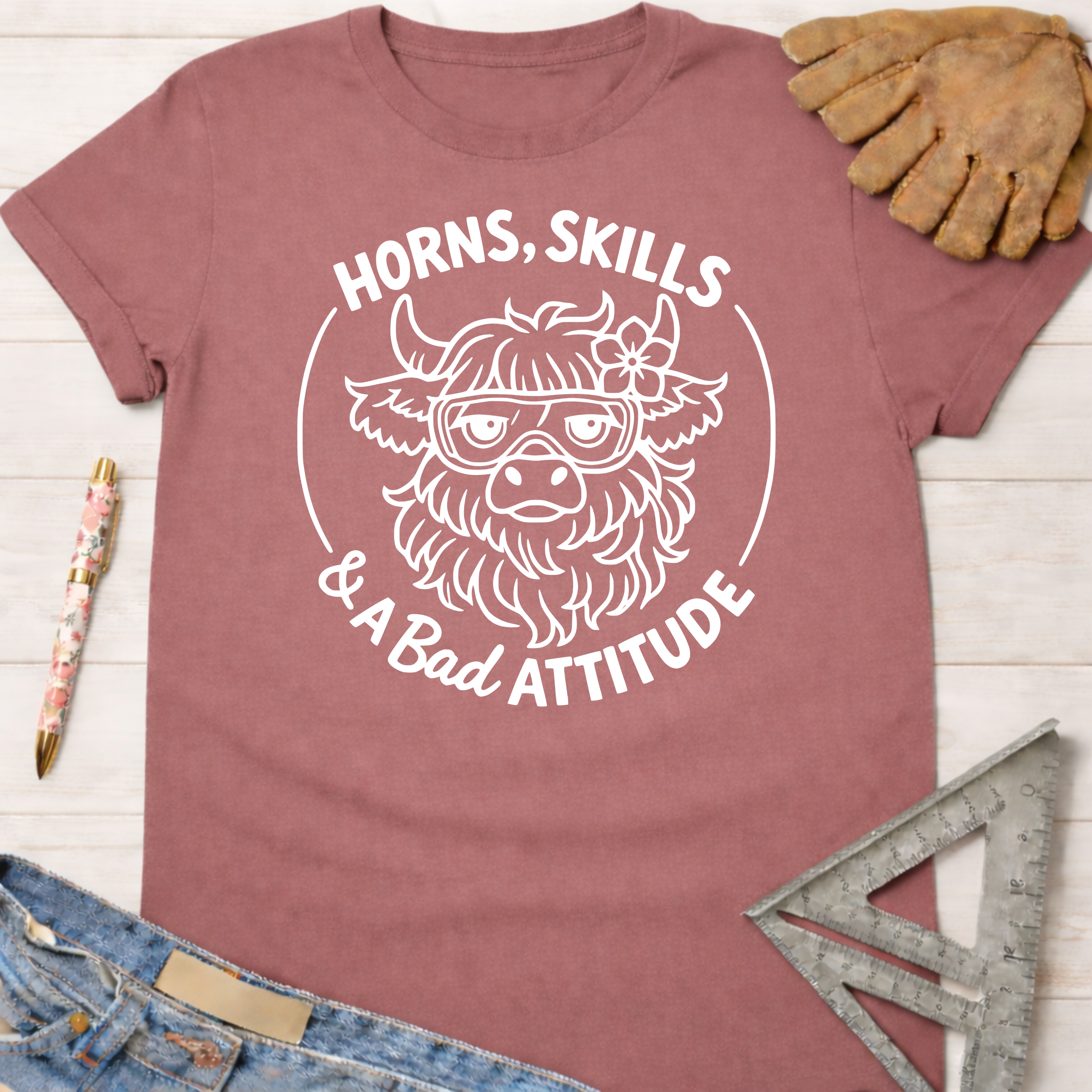 Women's funny woodworking graphic tee shirt for female woodworkers, makers, and DIY builders — Sawdust & Ponytails. Mauve t-shirt with a white graphic of a horned cow wearing goggles and a flower, text "Horns, Skills & A Bad Attitude.